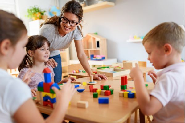 Teacher guiding young children as they play and learn with colorful building blocks at a table.
